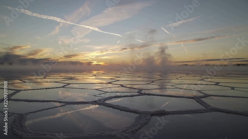Gliding low drone capturing polygonal saltpan reflections while sunrise warming water, raising mist