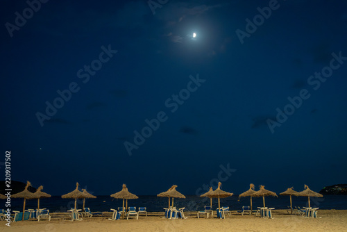 Empty sandy beach with thatched straw umbrellas under the moonlit night sky