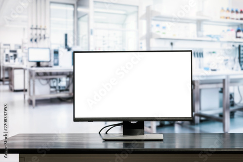 Blank desktop computer monitor on a dark table in a bright modern laboratory, with blurred scientific equipment and workspace in the background.