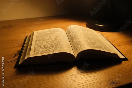 Open holy book bible on wooden table under warm desk lamp light focus