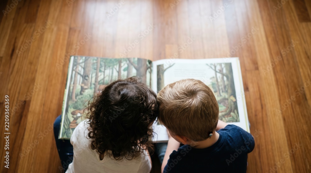 Fototapeta premium Two toddlers reading a large open picture book together on a wooden floor, capturing childhood curiosity and early literacy for educational marketing and preschool storytelling.