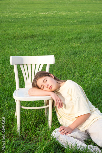 Young woman sitting alone on a white wooden chair in a lush green meadow.