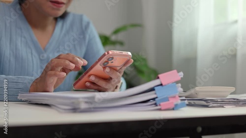 Female office staff using smartphone while reviewing company file folders handling client correspondence and checking daily work schedule at desk in bright modern office room