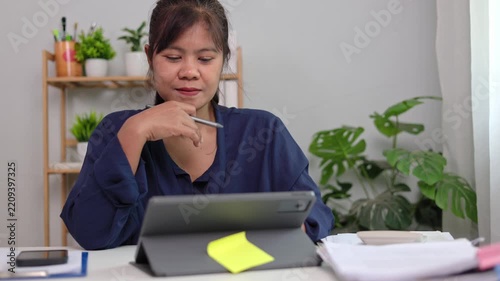 Asian female manager observing digital report displayed on tablet stand while making notes during online video conference in quiet well lit office background area room space