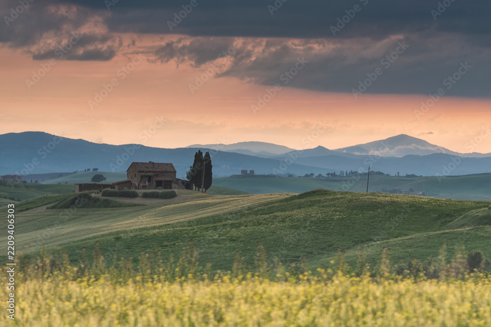 Obraz premium Traditional stone farmhouse standing on rolling green hills in Tuscany at sunset, framed by distant mountains and a dramatic evening sky. Ideal for travel and agritourism concepts.
