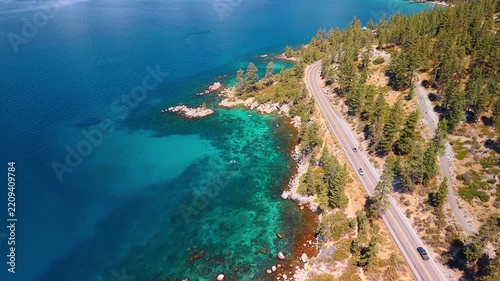 Lakeside highway winding through forest above Lake Tahoe. Cars follow a curving road between conifers and turquoise Lake Tahoe shallows beneath a wooded slope.