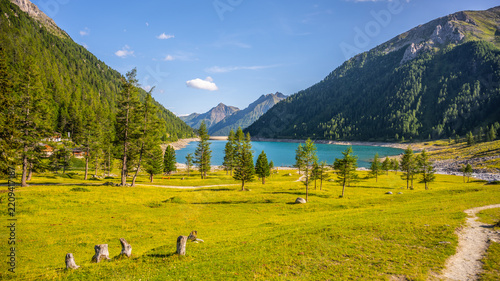 Visitors enjoy the beauty of Lake Neves located at the end of Molini Valley in the Zillertal Alps, Italy. Green grass and trees surround the lake under a clear blue sky.