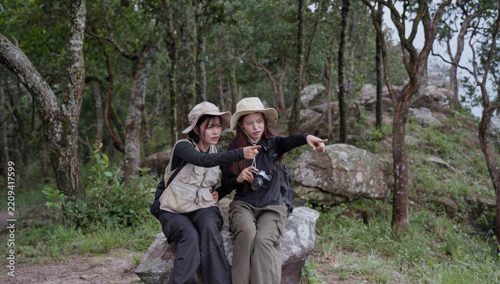 Fototapeta premium Two women hikers rest on a large rock in a green forest, pointing toward a distant view while carrying a camera and backpacks during a shared outdoor travel and nature adventure together.