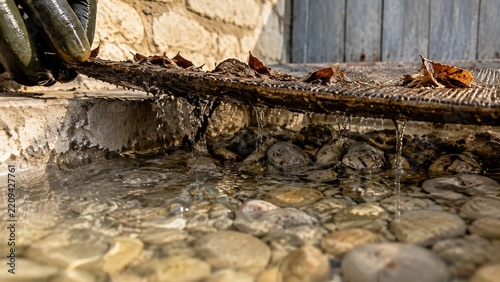 Water drips from a natural debris overhang into shallow flowing water over smooth river rocks, showing droplets forming and falling steadily into the basin