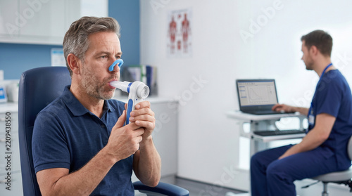 Mature man performing lung function test focused on health with calm expression during medical examination in clinic room at hospital