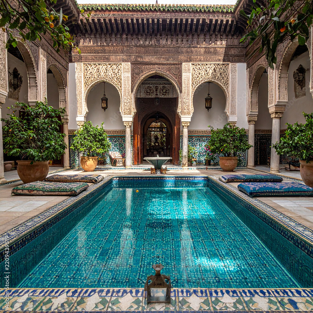 Obraz premium A serene Moroccan riad courtyard featuring a sparkling turquoise tiled pool, elegant arched architecture, and lush potted orange trees. Nobody is present in this tranquil, sunlit oasis of calm.