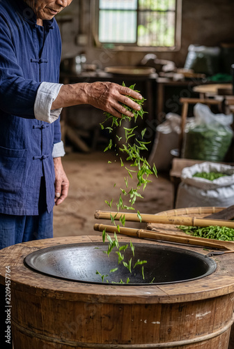 Senior Asian tea master dropping fresh green tea leaves into traditional wooden roasting tub.