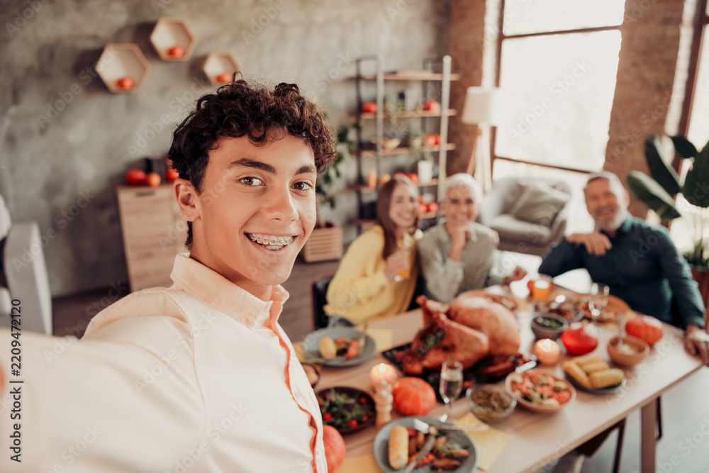 Obraz premium Teen boy taking a family Thanksgiving selfie at a holiday dinner table with relatives smiling, festive turkey, and warm home celebration
