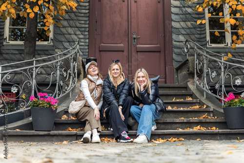 Three smiling women sit on stone steps before historic doorway during autumn trip in Harz, Germany. Candid travel portrait shows friendship, tourism, leisure and warm outdoor seasonal atmosphere
