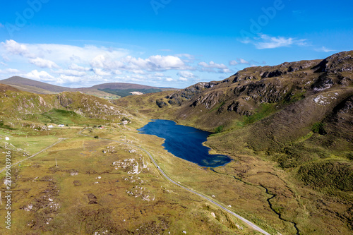 Aerial view of An Port lake surrounded by rugged mountains, green hills, and rocky terrain with a winding road in the valley in Ardara, County Donegal, Ireland.