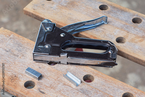 Heavy duty steel staple gun with metal staples on wooden workbench. Furniture upholstery, carpentry fastening, DIY home repair, construction tacker tool and woodworking concept.