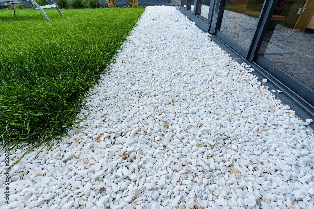 Fototapeta premium Strip of white decorative gravel between dark glass patio doors and lush green lawn in modern garden landscaping walkway