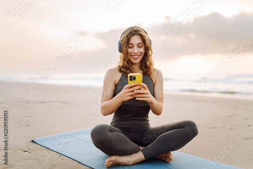Woman in sportswear sits on yoga mat on beach, wearing wireless headphones and holding phone. Athletic woman warms up and listens to music, enjoying seascape. Concept of enjoyment and relaxation.