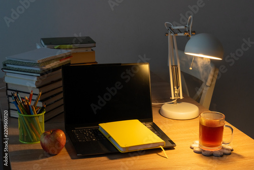 Cozy study desk with dark laptop screen, yellow notebook, books, colored pencils, apple and hot tea by a lamp. Concept of evening homework, exam preparation and healthy study breaks at home.