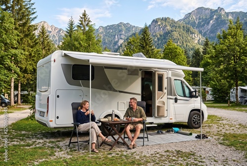 Two people are sitting at a table outside a camper. They are enjoying food and drinks at a campsite in the mountains. Trees surround the area