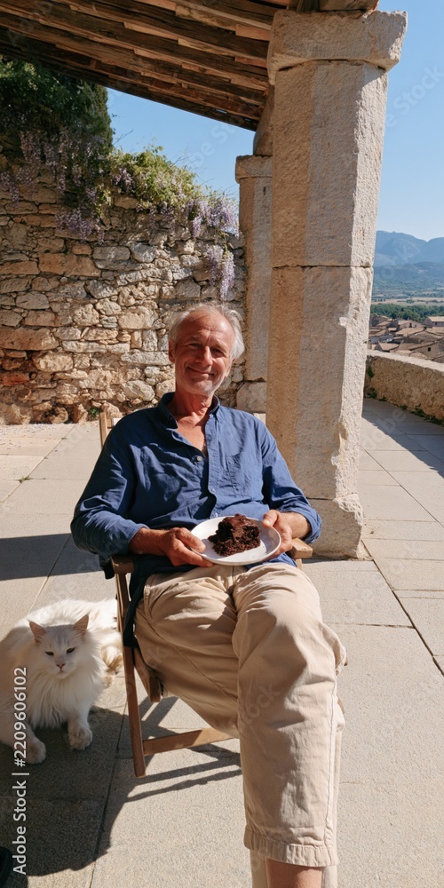 Fototapeta premium Older man holding plate of cake on sunny stone terrace with white cat for relaxed travel background and warm vacation lifestyle scene
