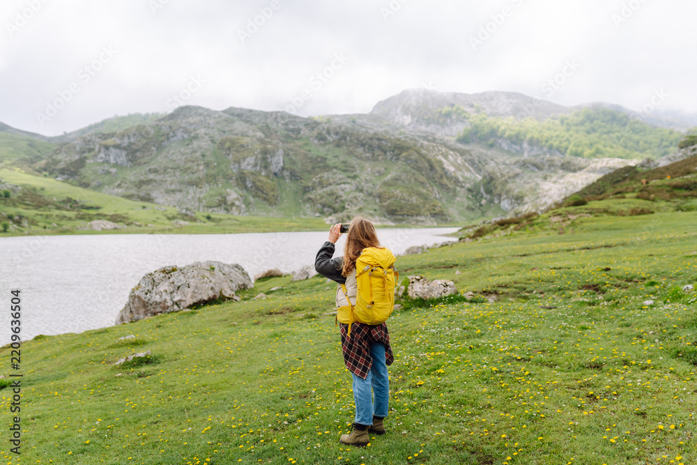 Obraz premium Female hiker with a yellow backpack uses her phone to photograph a mountain-ringed lake. She is enjoying nature and holding her smartphone on a cloudy day. Nature and adventure concept.
