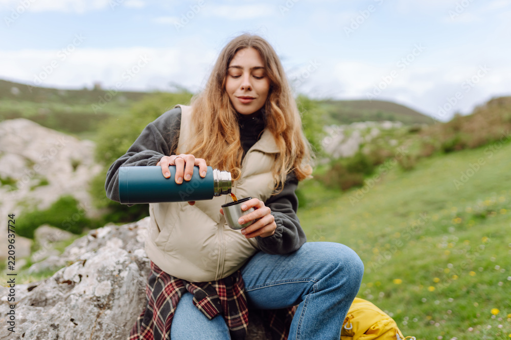 Naklejka premium Female hiker with backpack sits on large rock, pouring tea from thermos against picturesque mountain backdrop. Woman enjoys a rest and drinks a hot beverage while hiking. Adventure concepts.