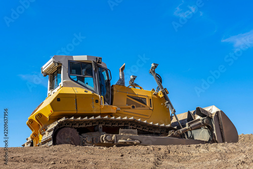 Massive, bright yellow bulldozer at construction site with tracks embedded in a mound of rich, brown soil. A clear blue sky creating a contrasty background.