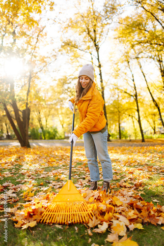 Woman in yellow jacket rakes leaves in park filled with colorful autumn foliage. Female volunteer clears away fallen leaves with yellow rake on autumn day. Concept of cleanup, ecology.