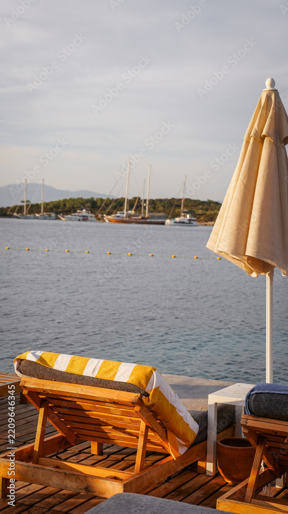 Fototapeta premium Wooden sun loungers with yellow striped towels on a seaside deck overlooking anchored boats
