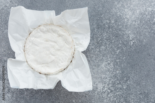 Round brie cheese wrapped in white paper on a grey surface