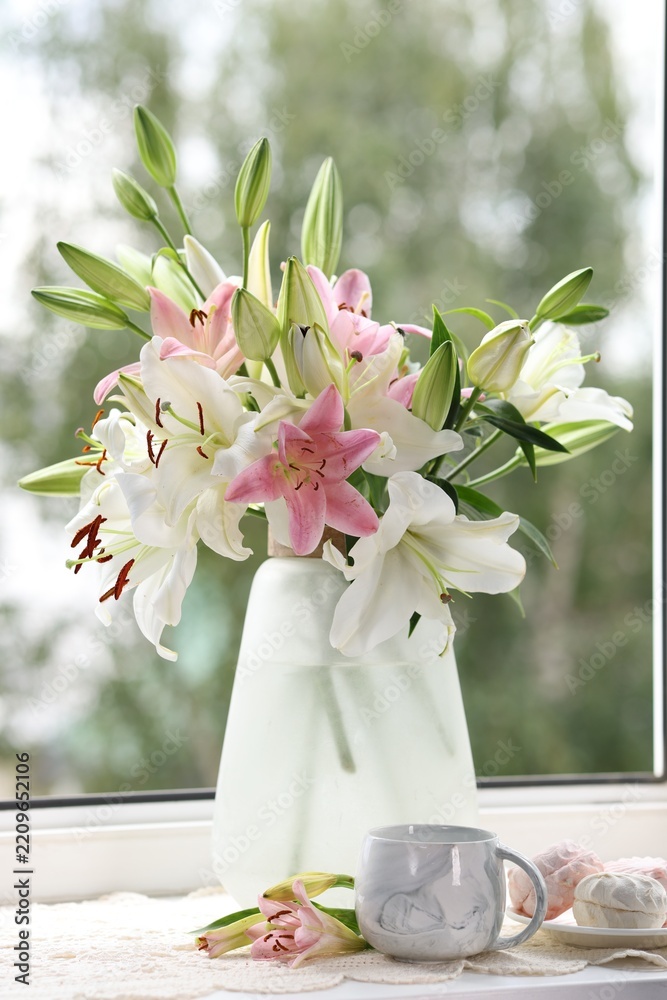Fototapeta premium Beautiful lily flowers in vase, zephyrs and cup of drink on window sill indoors