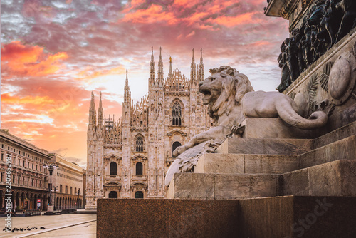 Scenic panoramic view of Milan cathedral and monument stone lion at sunrise with dramatic clouds and empty square