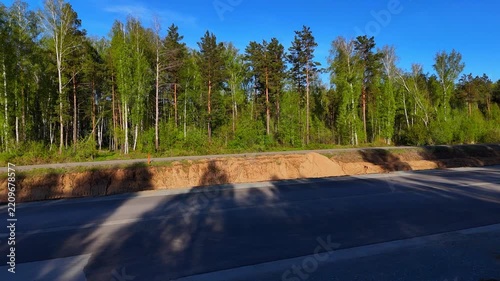 A sideways view to the right with a smooth left pan over a newly paved road surrounded by a pine forest, shot on a sunny summer evening. Eastern Bypass, Novosibirsk, Russia.