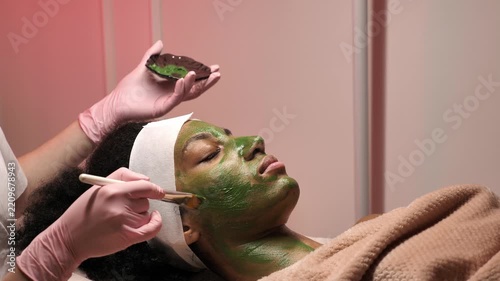 Cosmetologist applying green clay face mask with brush on female client face in spa salon. Skin care treatment, organic cosmetic facial procedure, rejuvenation, facial care and wellness concept