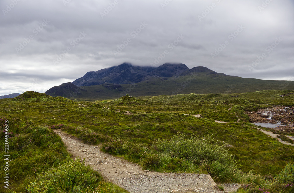 Fototapeta premium Hiking trail leading through green moorland towards the dramatic dark peaks of the Black Cuillin mountains on the Isle of Skye, Scotland.