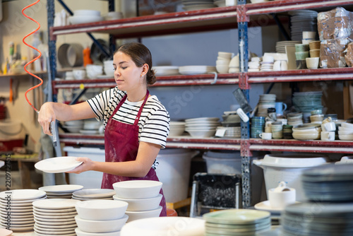 Young woman worker in uniform paints blanks of ceramic products at factory