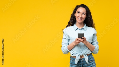 Smiling Young Woman With Dark Hair And Light Skin Holding Smartphone While Looking Upjoyfully At Studio Background. She Engaged In Communication, Expressing Positivity And Connection.