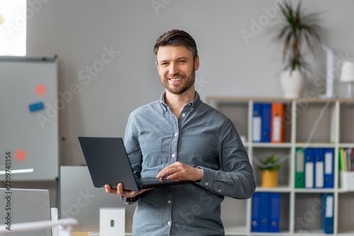 Smiling Man Standing With Laptop In Office Setting, Engaged In Work Activities. Professional Male In Mid-30s Wearing Casual Shirt, Represents Modern Business Concept.