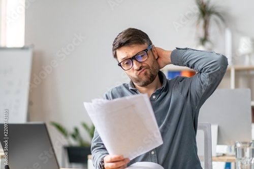 Worried Caucasian Man In His 30s Sitting At Desk In Modern Office, Reading Important Documents With Concerned Expression. Feeling Overwhelmed While Working On Business Task At Workplace.