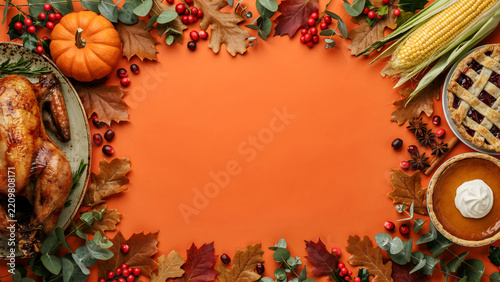 Thanksgiving dinner table setting with roasted turkey, pumpkin, corn, and pies on orange background with autumn leaves and berries, top view with copy space