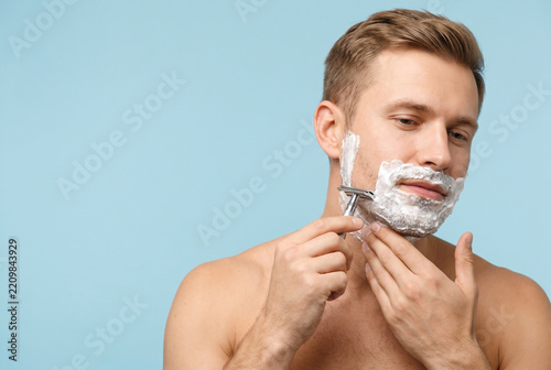Shirtless man applying shaving cream and using a double edge razor on his cheek, morning grooming routine and personal hygiene isolated on light blue