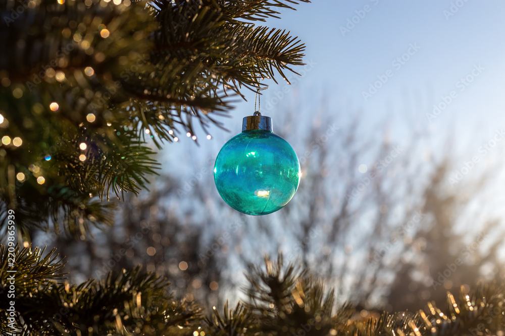 Fototapeta premium Turquoise Christmas bauble hanging from spruce branch outdoors. Christmas holiday decoration with golden reflections. Seasonal celebration and winter greeting.