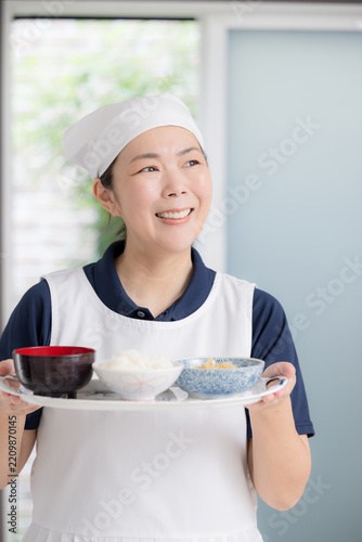 高齢者の自宅で三角巾をつけ食事を用意する笑顔の女性スタッフ、家事代行、料理代行、訪問介護、食事支援、配膳イメージ笑顔