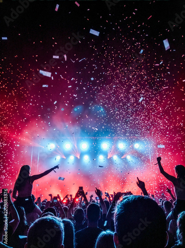 Concert crowd silhouettes cheering with falling confetti under red and blue stage lights and smoke