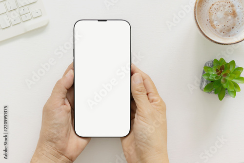 Top view of hand holding modern smartphone with blank white screen template and keyboard and small green plant, coffee on white desk. Concept for marketing, website, branding, social.