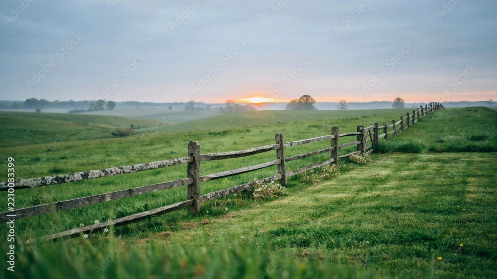 Fototapeta premium A weathered wooden split rail fence stretches across a green misty meadow at sunrise hour with rolling meadow landscape divides grassy rolling