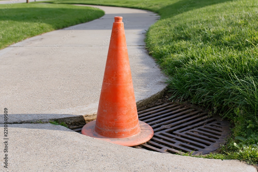 Obraz premium The Orange Traffic Cone Guarding a Street Drain on a Sunny Sidewalk