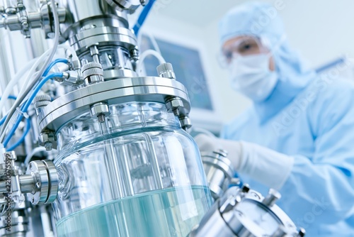 Closeup of stainless steel bioreactor vessel filled with pale green fermentation liquid surrounded by tubing and pressure fittings, researcher in full sterile lab suit and face mask observing