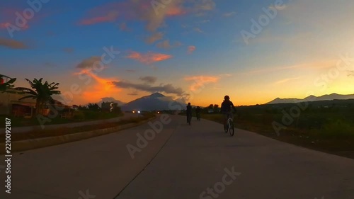 Cyclists Riding Bicycles on a Rural Road at Sunset with Mountain Silhouette and Colorful Twilight Sky